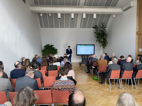 zum Auftaktevent im Dachfoyer des Haus-, Hof- und Staatsarchivs steht Richard Lein vor dem Publikum sitzend von hinten und spricht zu ihnen, neben ihm ein screen mit Präsentation